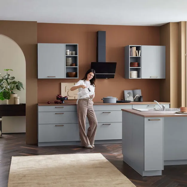 Moderne Bauformat Küche bei Küchenstudio Depping mit grauen Fronten und warmer Akzentwand, Frau beim Zubereiten in offener Wohnküche